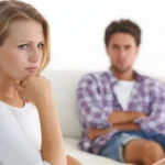A-woman-sitting-on-a-couch-with-a-thoughtful-expression-a-man-in-the-background-blurred-and-sitting-with-arms-crossed-1-1024x576